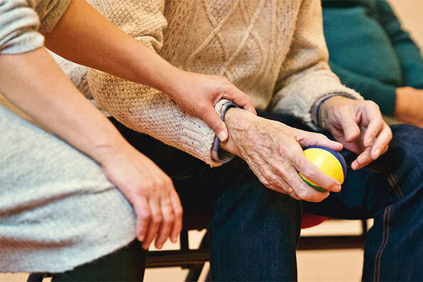 Three people sitting at assisted living facility while young hand touches old man's wrist that is holding a small ball.